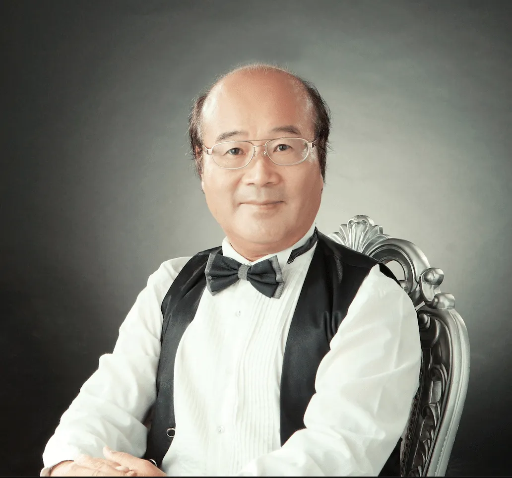 Portrait of a man wearing a tuxedo with a bow tie, seated elegantly in a decorative chair, showcasing a confident smile against a dark background.