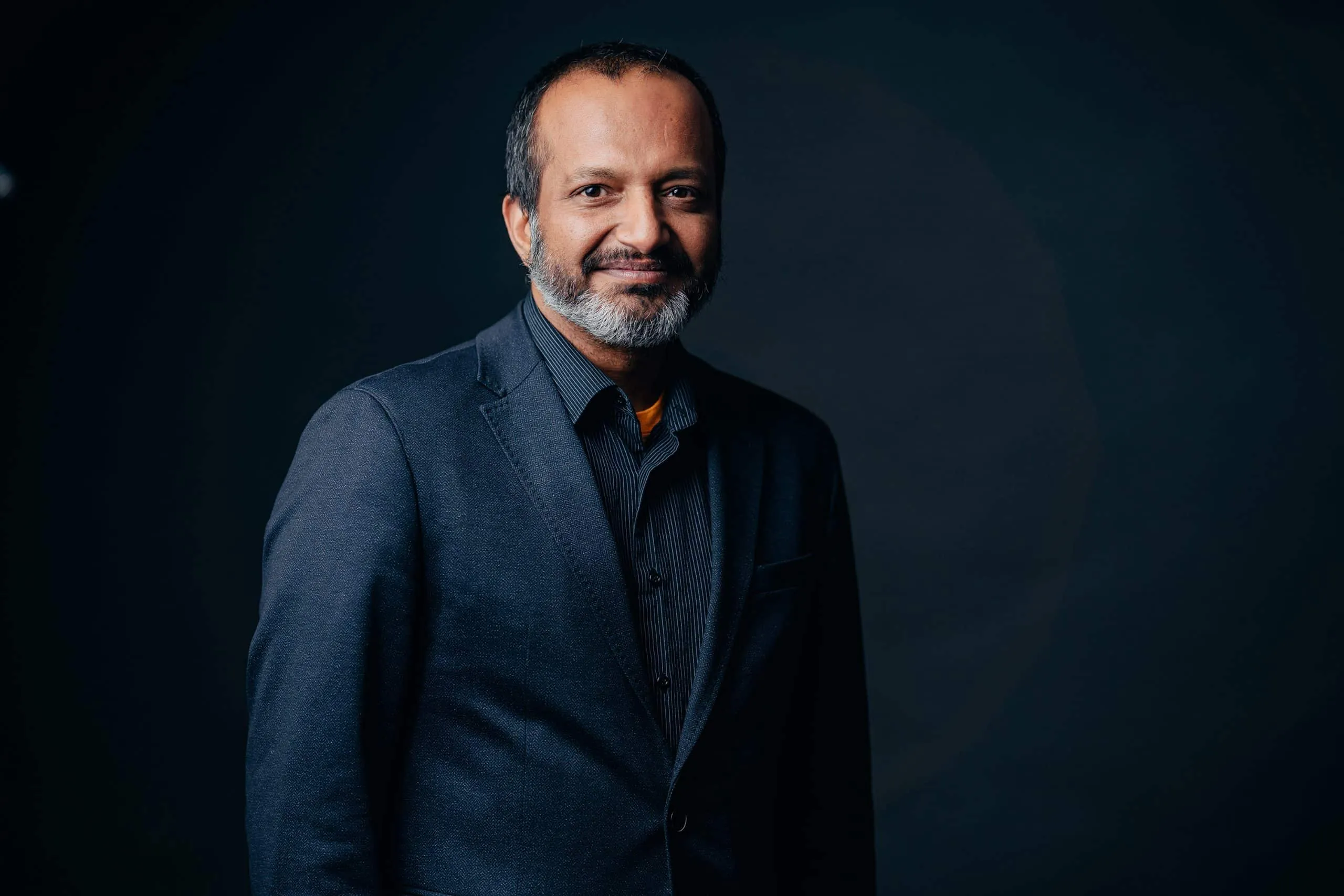 Professional portrait of a man with a beard, wearing a dark suit and button-up shirt, standing against a dark background, conveying a confident and approachable demeanor.