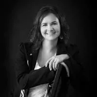 Portrait of a smiling woman with curly hair, wearing a black blazer and leaning on a chair, set against a dark background.