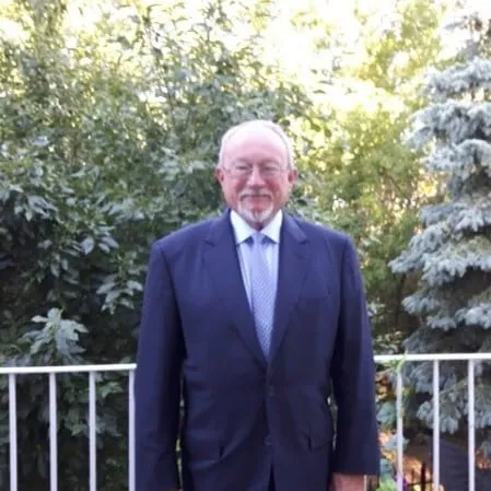 Elderly man in a dark suit standing outdoors in front of lush greenery and a railing.