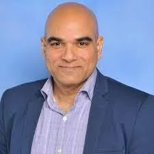Professional headshot of a bald man in a blue blazer and light purple shirt, smiling against a light blue background.