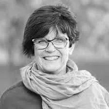 Smiling woman with short dark hair, wearing glasses and a light scarf, against a blurred background.