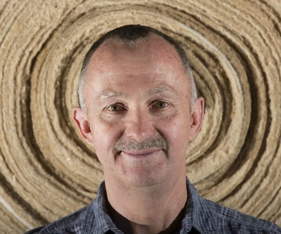 Smiling middle-aged man with a mustache, wearing a checked shirt, poses in front of a textured circular background. The image highlights his friendly expression and the natural tones of the backdrop.