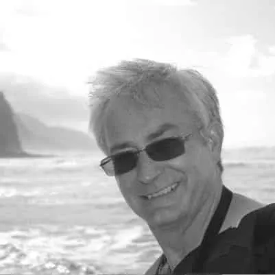 Smiling man with gray hair and sunglasses against a scenic coastal backdrop, capturing a joyful moment by the ocean.