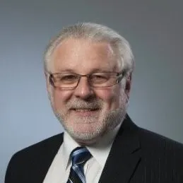 Professional headshot of a middle-aged man with gray hair and glasses, wearing a dark suit and striped tie, against a neutral background.
