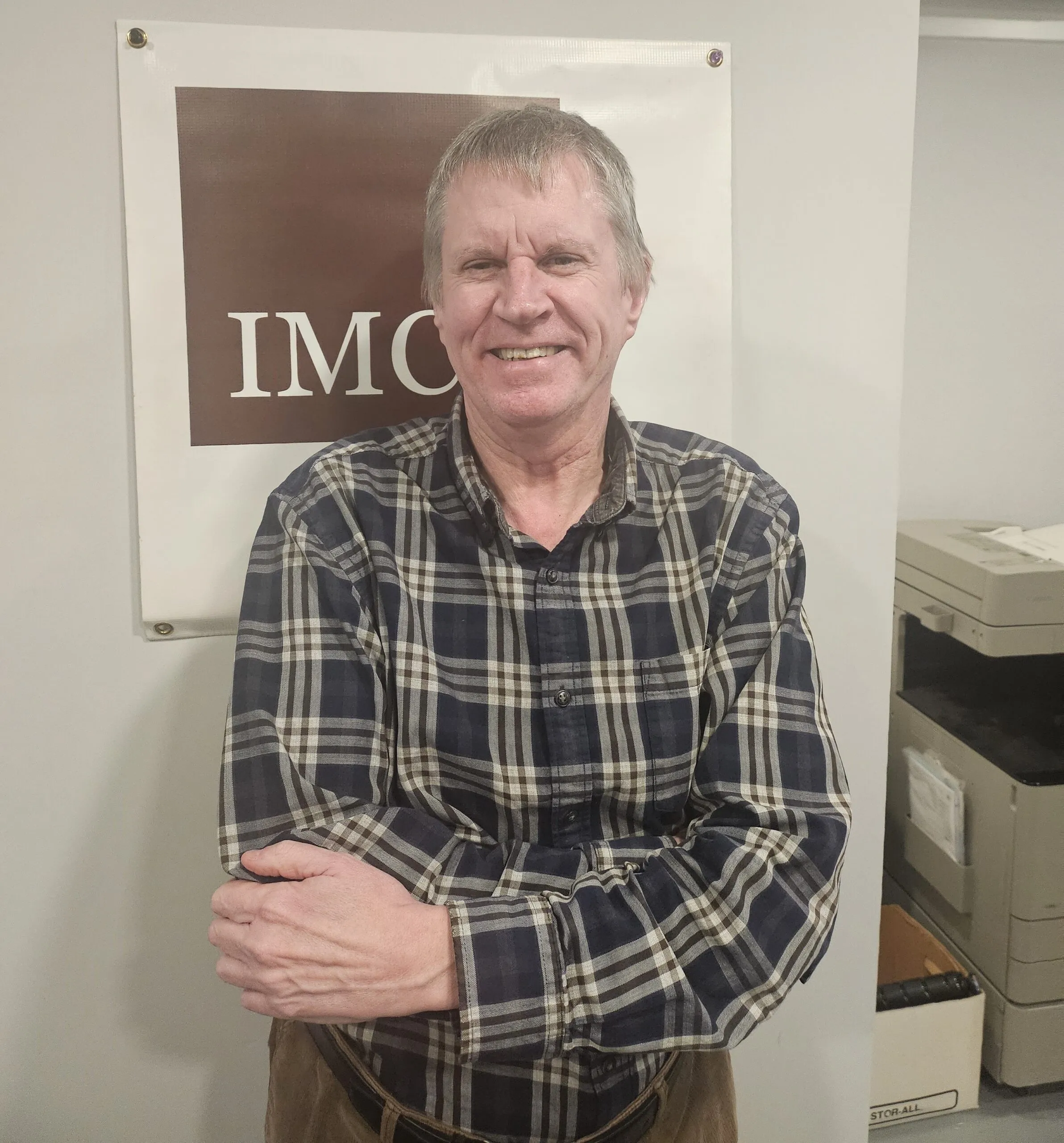 Smiling man with gray hair wearing a plaid shirt stands with crossed arms in front of an IMC sign, with office equipment visible in the background.