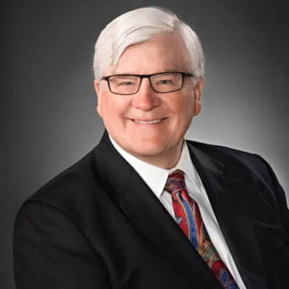 Professional headshot of a smiling older man with gray hair, wearing glasses and a black suit jacket over a white shirt, paired with a colorful tie, set against a dark background.