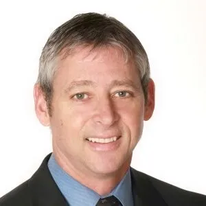 Professional headshot of a smiling man with short gray hair, wearing a black suit and light blue shirt, suitable for business or corporate profiles.
