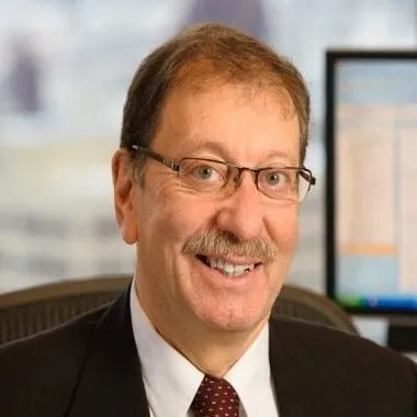 Smiling professional man with glasses, wearing a suit and tie, seated in an office environment with a computer screen in the background.