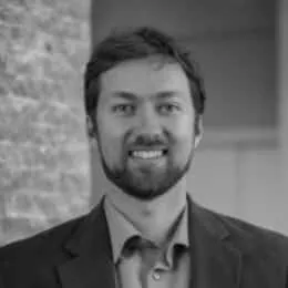 Professional headshot of a smiling man with a beard, wearing a suit and collared shirt, standing against a textured wall background.