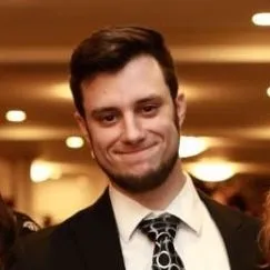 Young man in a formal suit and tie smiling at a social event, with a softly lit background.
