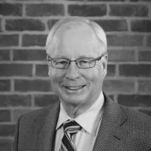 Professional portrait of a smiling older man wearing glasses and a patterned suit, set against a brick wall background.