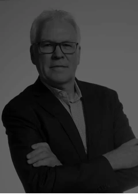 Professional headshot of a middle-aged man wearing glasses and a dark blazer, with arms crossed and a confident expression, set against a neutral background. Ideal for business or professional profiles.