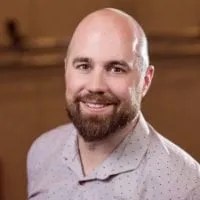 Smiling man with a beard wearing a light-colored patterned shirt, posing in a warm-toned indoor setting.