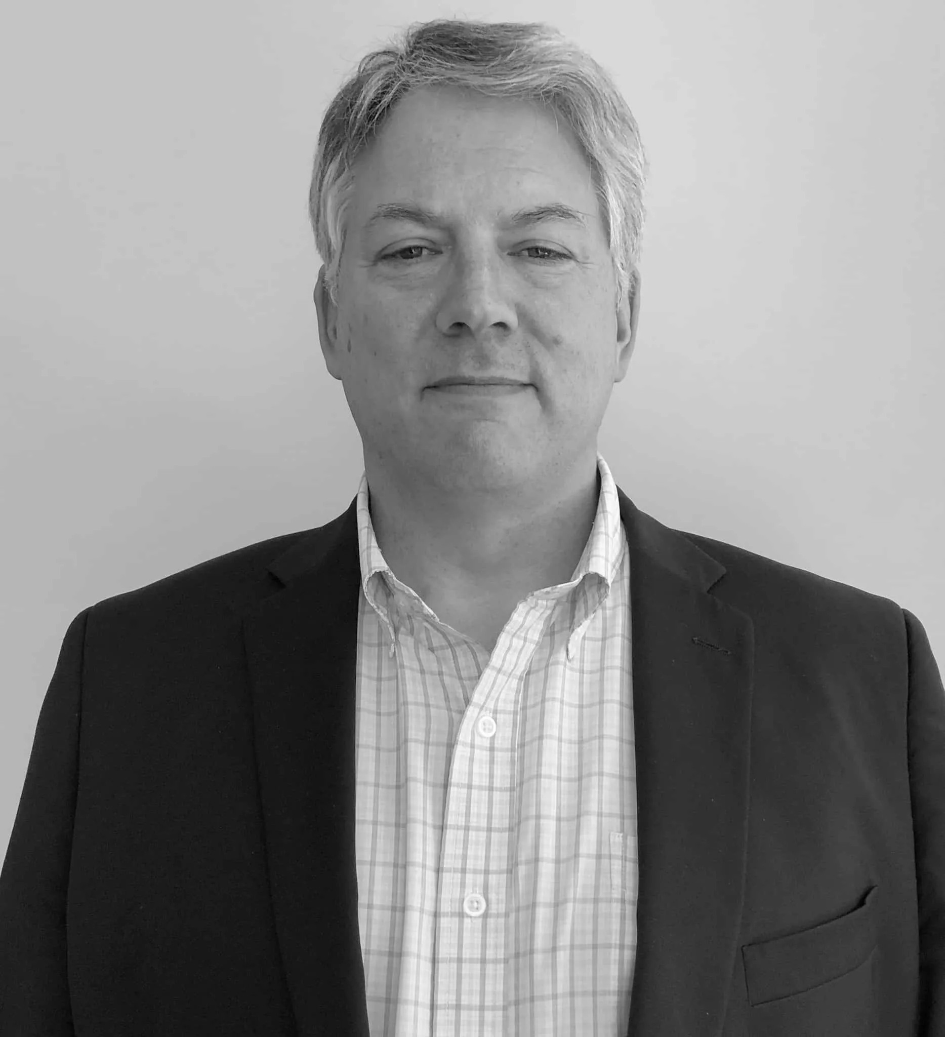 Professional headshot of a man with gray hair wearing a black blazer and a plaid shirt, standing against a neutral background.