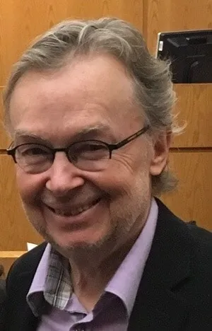 Smiling man with glasses and gray hair, wearing a black blazer over a purple shirt, standing in a courtroom setting with wooden paneling in the background.
