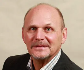 Professional headshot of a middle-aged man with short hair and blue eyes, wearing a dark blazer over a checkered shirt, smiling against a neutral background.
