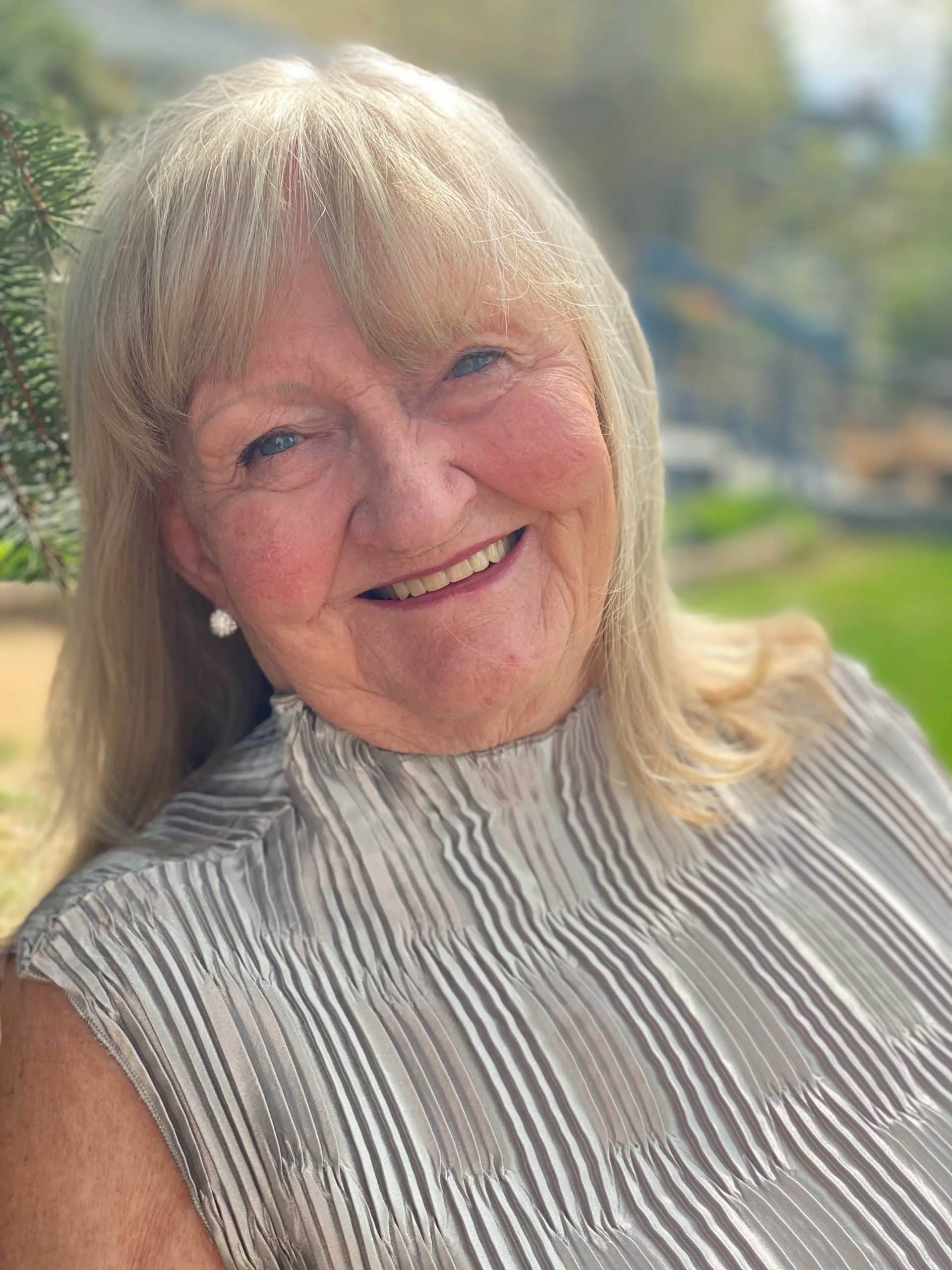Smiling elderly woman with long blonde hair wearing a pleated silver top, posing outdoors with greenery in the background.