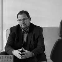 Business professional seated in a modern office, wearing a suit and glasses, with a contemplative expression.