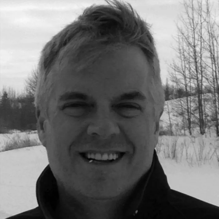 Smiling man with short, light-colored hair wearing a dark jacket, standing outdoors in a snowy landscape with trees in the background.