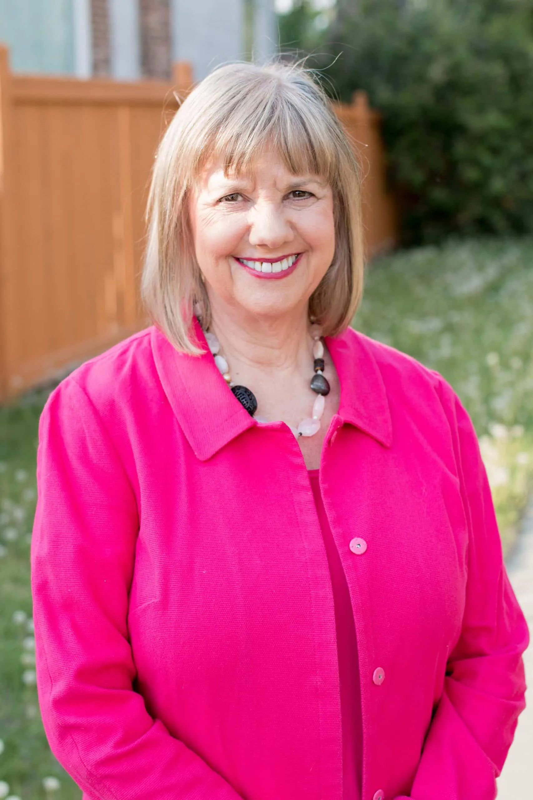 Smiling woman wearing a bright pink jacket and necklace, standing outdoors in a garden setting with a wooden fence in the background.