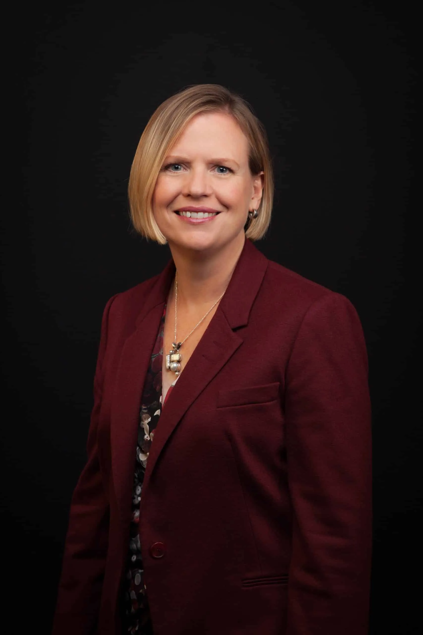 Professional portrait of a woman with short blonde hair, wearing a maroon blazer over a floral blouse, smiling against a black background. Ideal for business profiles or corporate presentations.