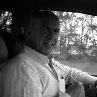 Smiling older man in a vehicle, wearing a light-colored shirt, with trees visible in the background, conveying a sense of enjoyment while driving.
