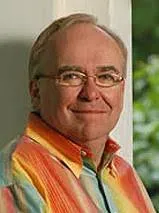 Portrait of a middle-aged man wearing a colorful, striped shirt, smiling softly while standing indoors near a window with greenery in the background.