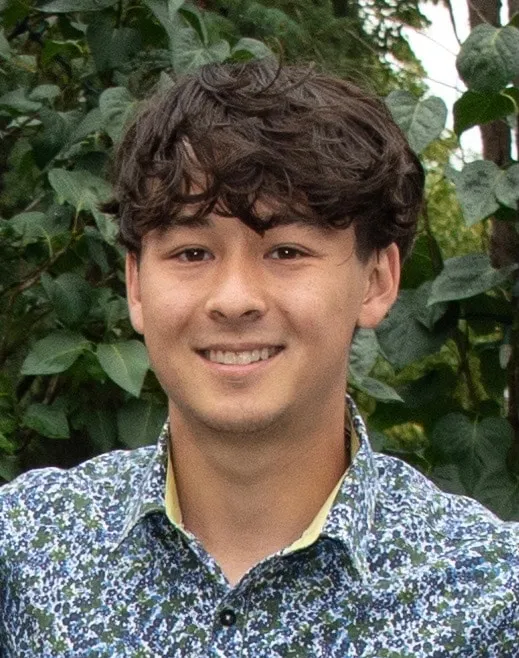 Smiling young man with curly hair wearing a floral patterned shirt, standing outdoors among green foliage.
