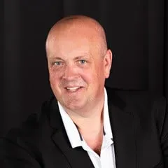 Smiling man in a black suit and white shirt, posing against a dark background, exuding confidence and professionalism.