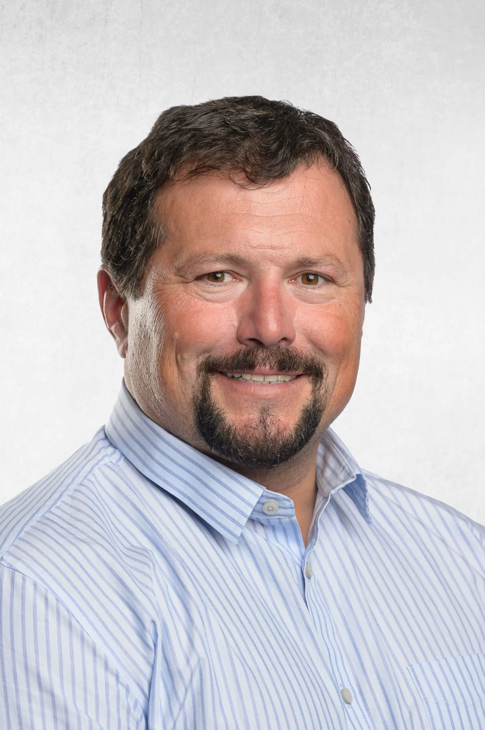 Portrait of a smiling man with short, dark hair and a beard, wearing a light blue striped shirt. The background is a soft gradient, enhancing the professional appearance of the image.