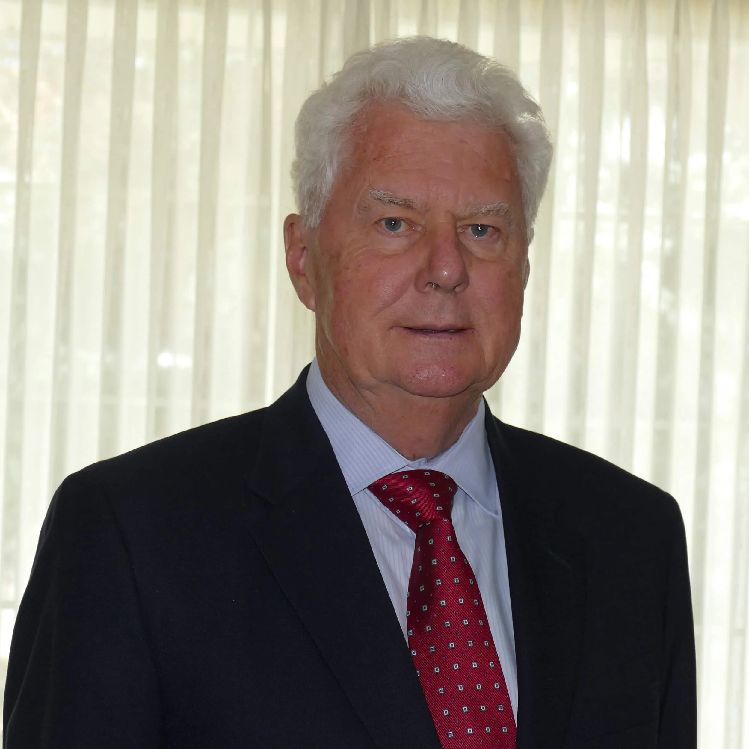 Portrait of a mature man with white hair, wearing a dark suit and red tie, standing in front of sheer curtains. The image conveys a professional and formal atmosphere, suitable for business or executive profiles.