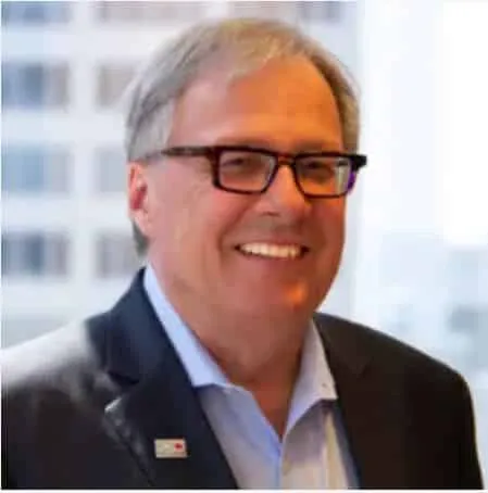 Smiling middle-aged man in a business suit with glasses, standing by a window in an office setting, showcasing a professional demeanor.
