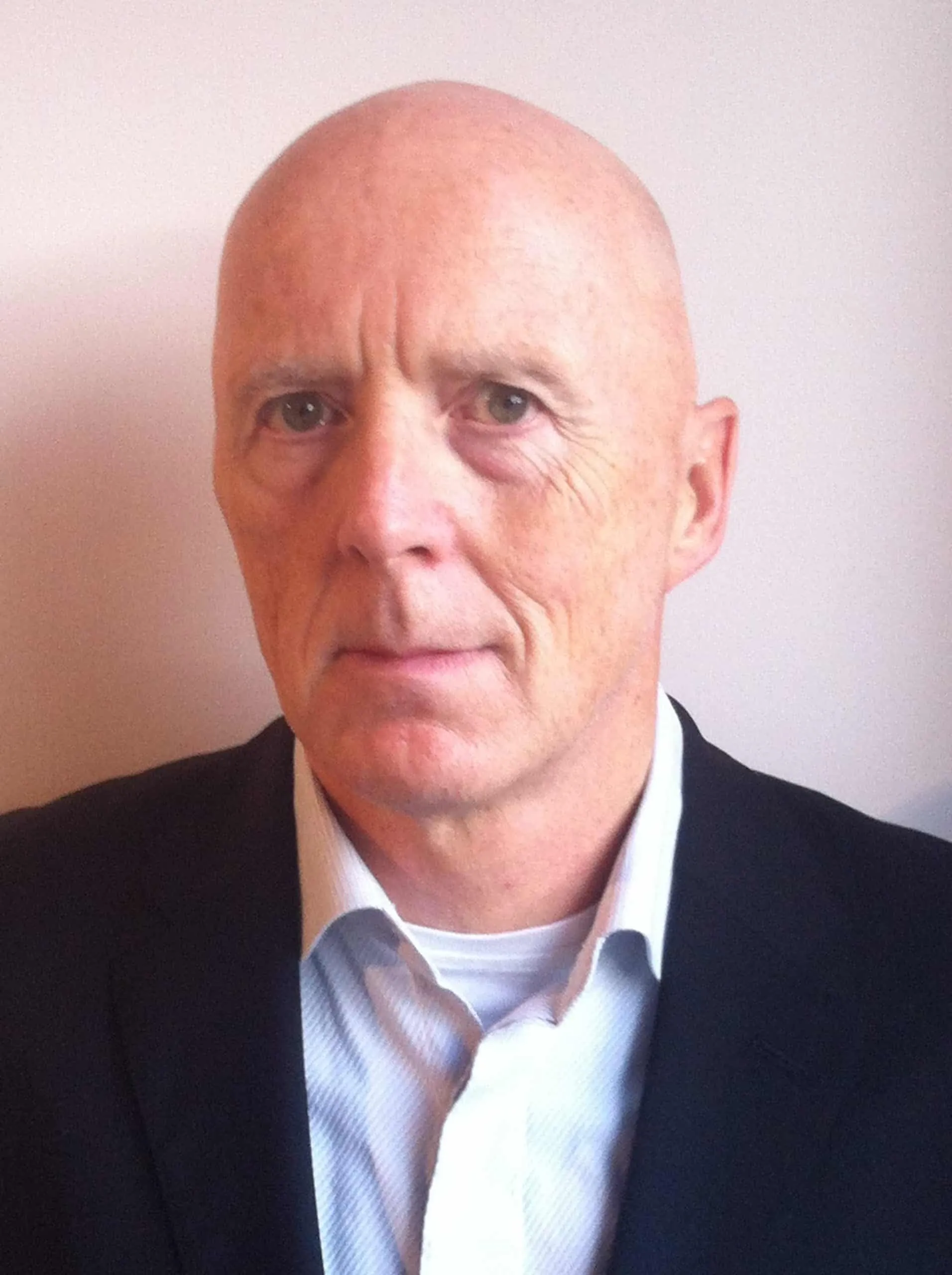 Portrait of a middle-aged man with a bald head, wearing a dark suit jacket over a light blue shirt, looking directly at the camera with a neutral expression against a plain background.