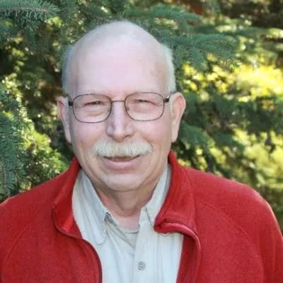 Smiling elderly man wearing glasses and a red jacket, standing outdoors in front of evergreen trees.