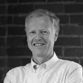 Smiling middle-aged man in a collared shirt, standing with arms crossed against a brick wall background.