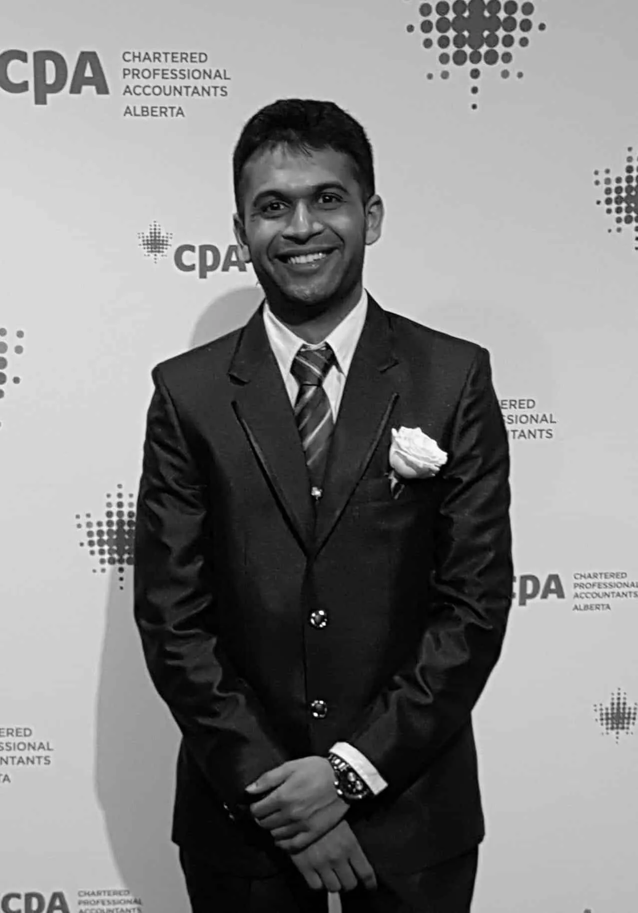 Young professional in a suit and tie with a white rose pinned to his lapel, smiling in front of a backdrop featuring the CPA (Chartered Professional Accountants) logo in Alberta.