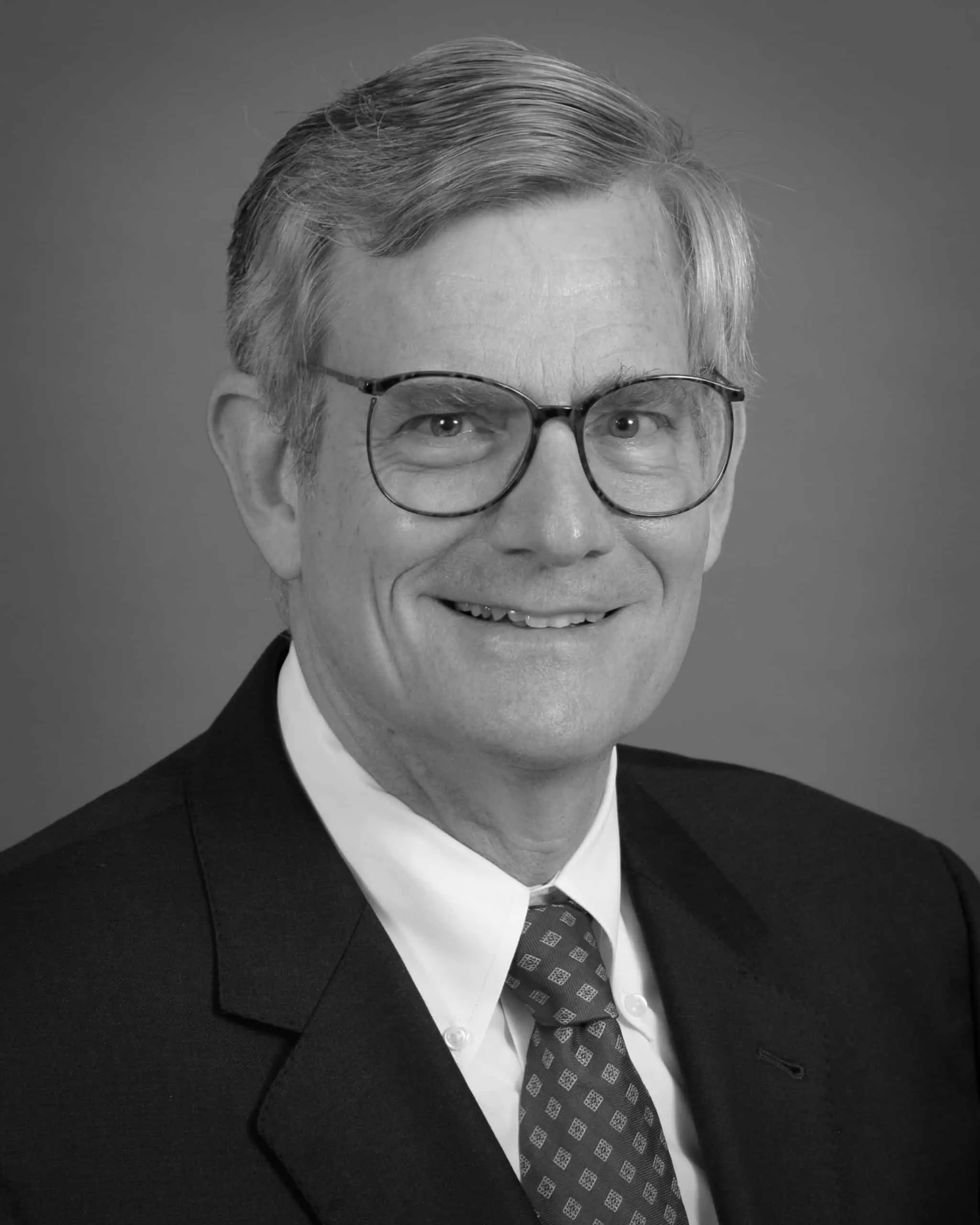 Portrait of a middle-aged man with gray hair, wearing glasses and a suit, smiling against a plain background.
