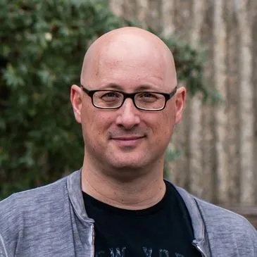 Bald man wearing glasses and a casual jacket, smiling outdoors with greenery in the background.