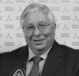 Man in a suit and glasses smiling at the Emerald Awards backdrop, showcasing a formal event atmosphere.