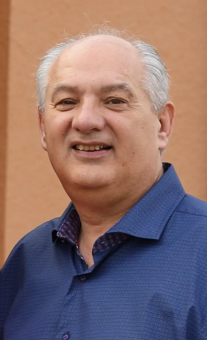 Smiling middle-aged man in a blue patterned shirt, standing outdoors against a warm-toned background. Ideal for business or personal profile imagery.