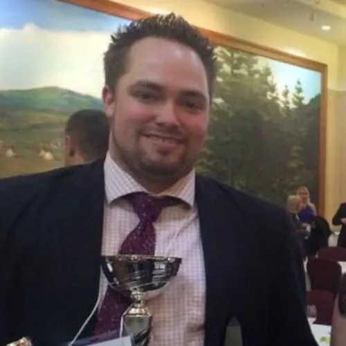 Man holding a trophy at an award ceremony, wearing a suit and tie, with a background featuring a scenic mural.