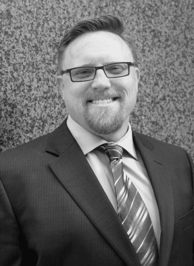 Professional headshot of a smiling man in a suit and tie, wearing glasses, against a textured gray background. The image conveys a confident and approachable demeanor, suitable for business or corporate use.