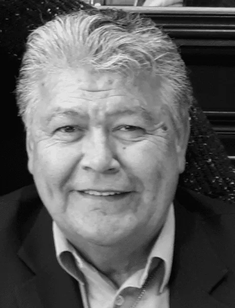 Smiling older man with gray hair, wearing a dark suit and light shirt, seated against a textured background.