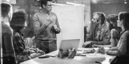 A diverse team of professionals engaged in a business meeting, with a speaker presenting ideas in front of a whiteboard, while others actively listen and take notes. A laptop and various documents are visible on the table, highlighting a collaborative work environment.