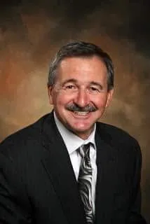 Professional headshot of a middle-aged man with a mustache, wearing a dark suit and patterned tie, smiling against a blurred, warm-toned background.