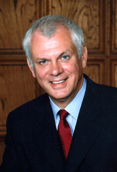Professional headshot of a smiling middle-aged man with gray hair, wearing a dark suit and a red tie, against a wooden background. Ideal for business or leadership-related content.