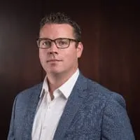 Professional portrait of a man wearing glasses and a blue blazer, standing against a dark wooden background, conveying confidence and professionalism.