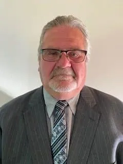 Businessman wearing glasses and a gray suit, standing against a neutral background. He has a white mustache and a patterned tie, exuding a professional appearance.