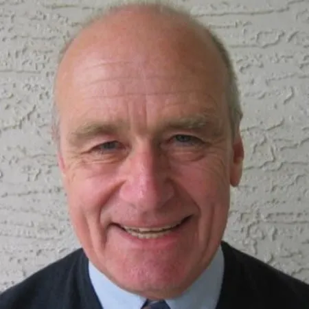 Smiling elderly man with gray hair, wearing a black sweater and light blue collared shirt, against a textured light-colored background.